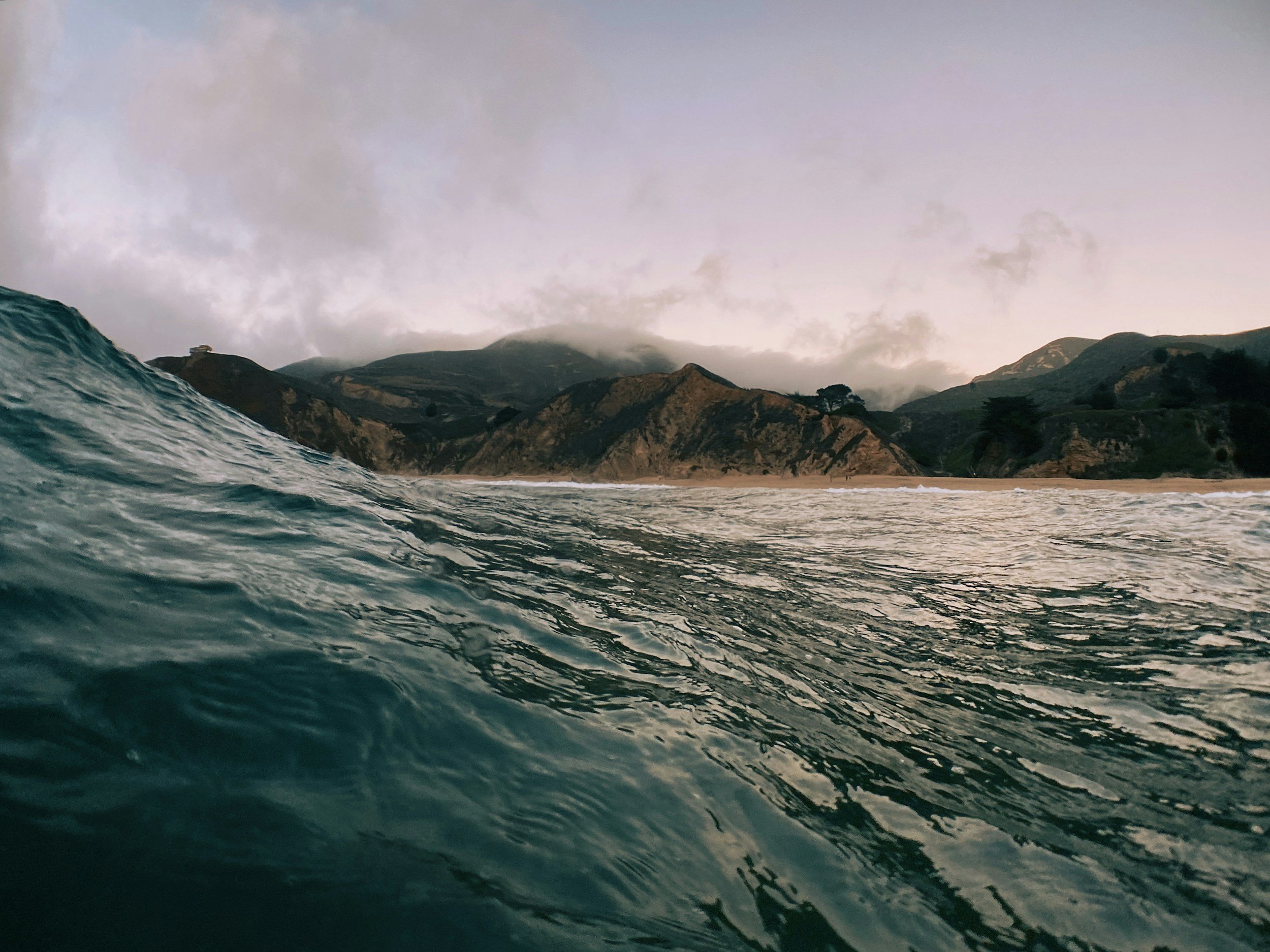 Ocean wave approaching sandy beach with hills and cloudy sky in the background.