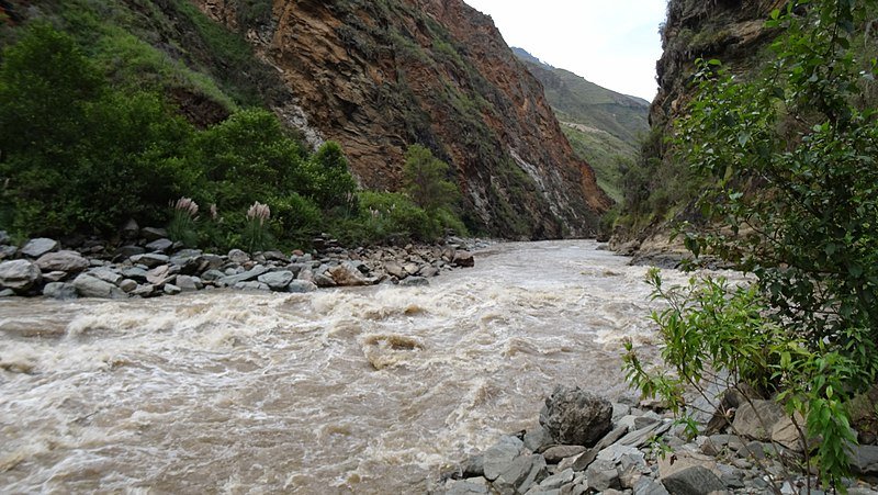 A rushing river flowing through a rocky canyon with green foliage on the sides and rugged brown cliffs in the background.