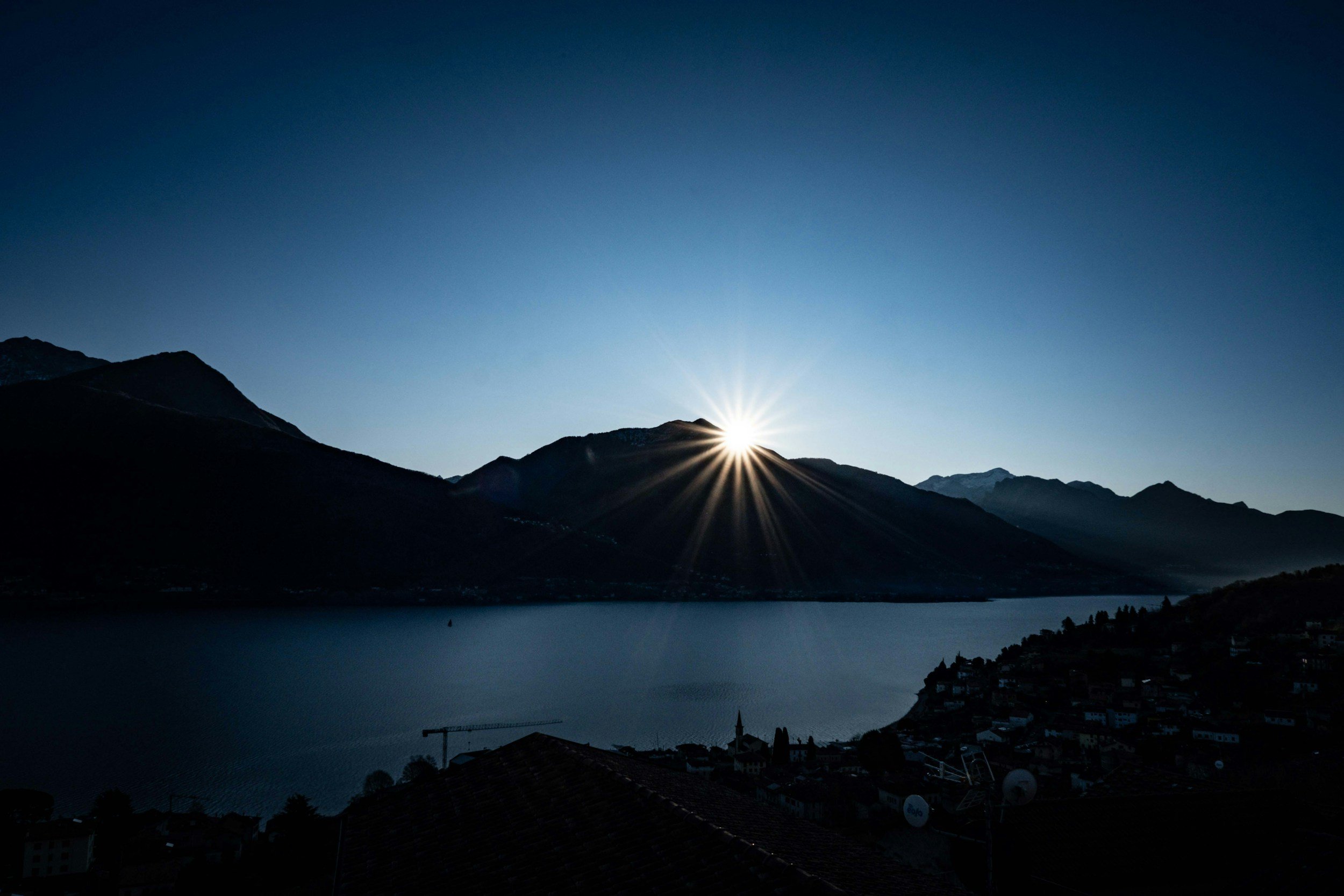 Sun rising behind mountains over a lake with rooftops in the foreground.